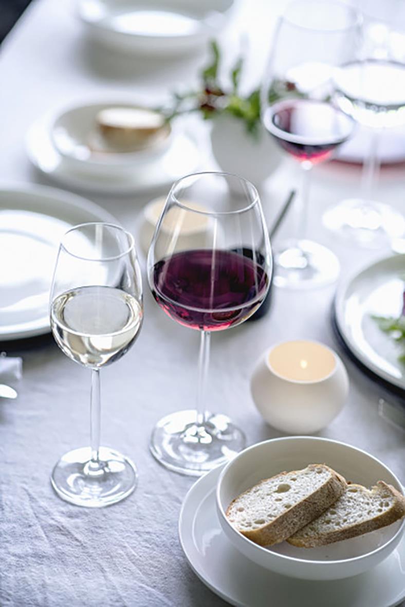 Vinglas av glas med röd och vitvin på dukat bord med tallrikar och ljus i bakgrunden