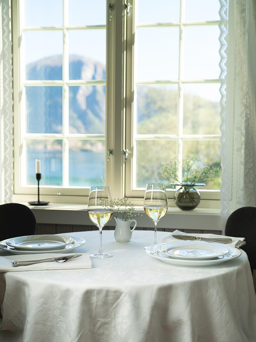 Vinglas på dukat bord med utsikt genom fönster mot berg och sjö, i ett ljust rum
