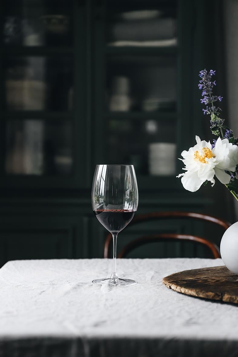Vinglas Riedel Veritas Cabernet/Merlot med rödvin på ett bord med blomsterarrangemang i bakgrunden