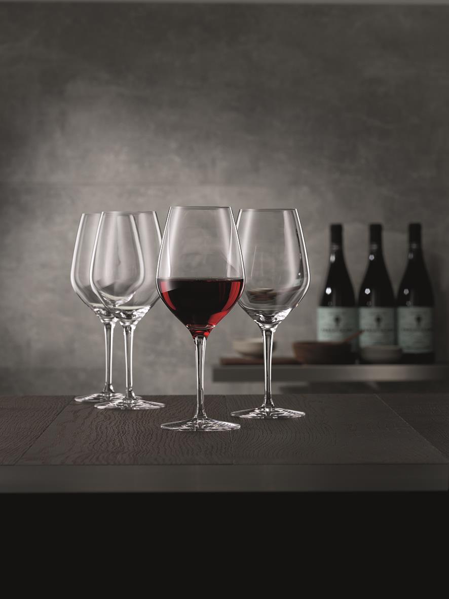 Tre vinglas, ett med rödvin, på mörk träbord mot mörk bakgrund med vinflaskor i bakgrunden