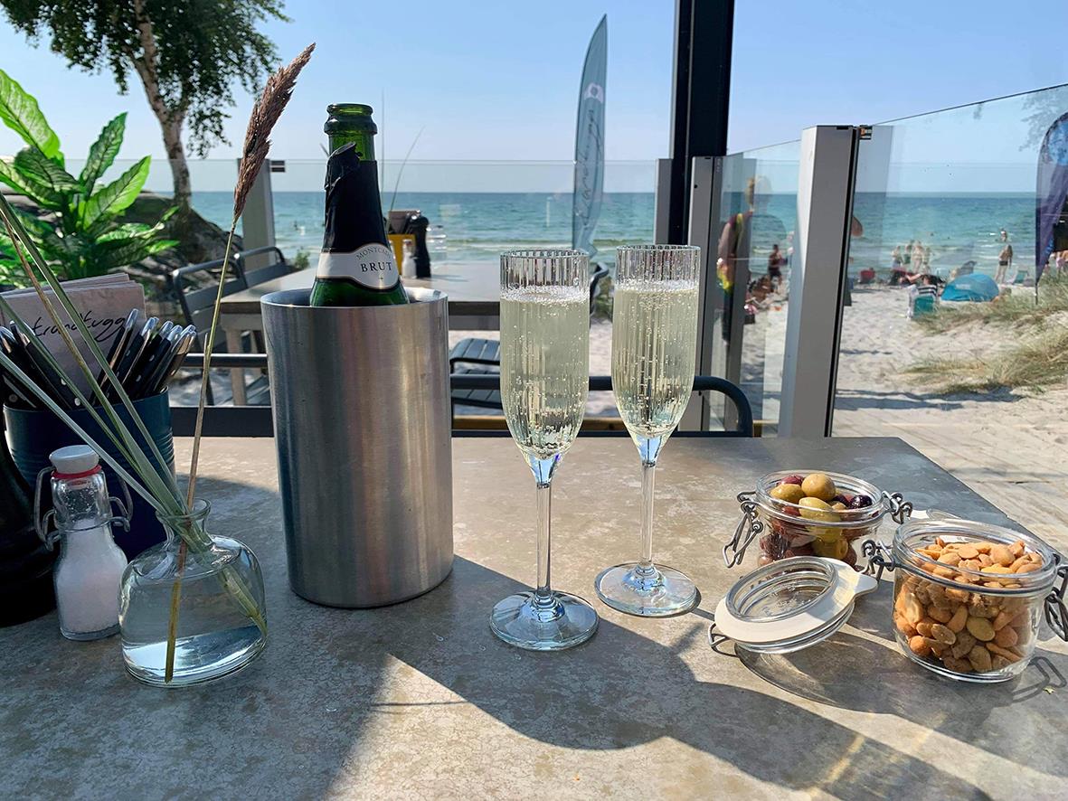 Champagneglas på bord vid strand med flaskor, snacks och utsikt över stranden och havet