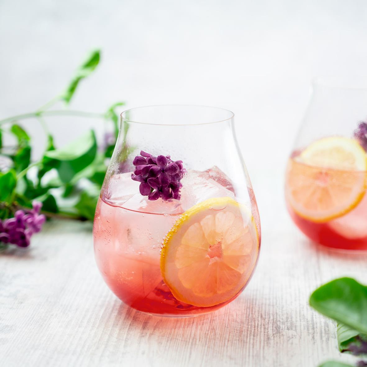 Gin & Tonic i Riedel-glas med citron och lavendel, på vit bakgrund med gröna blad