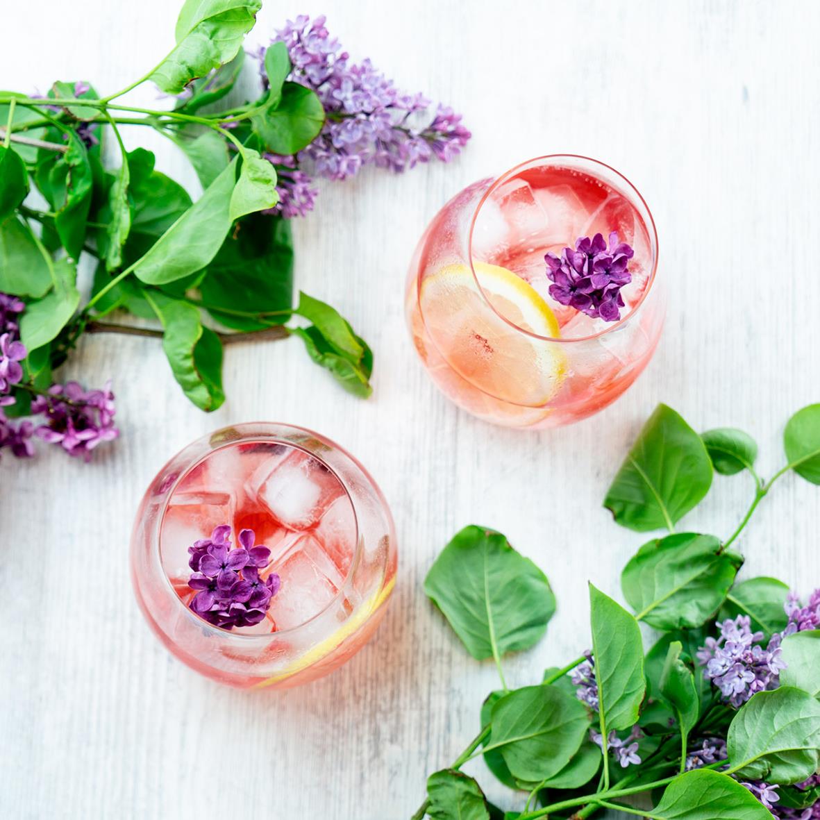 Två glas Gin & Tonic med citronskivor och blommor på ett vitt bord, omgivna av gröna blad och lila blommor