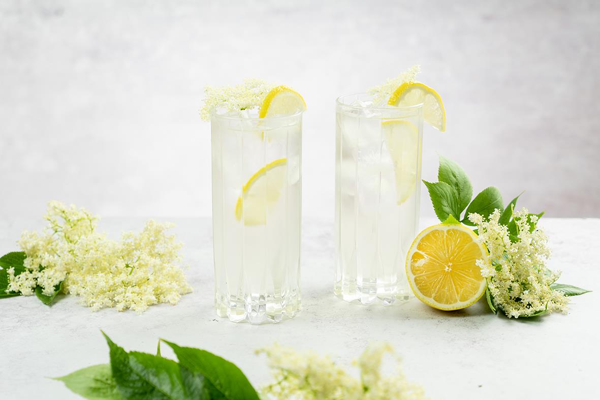 Två höga drinkglas med citron och is, omgivna av citron och vita blommor