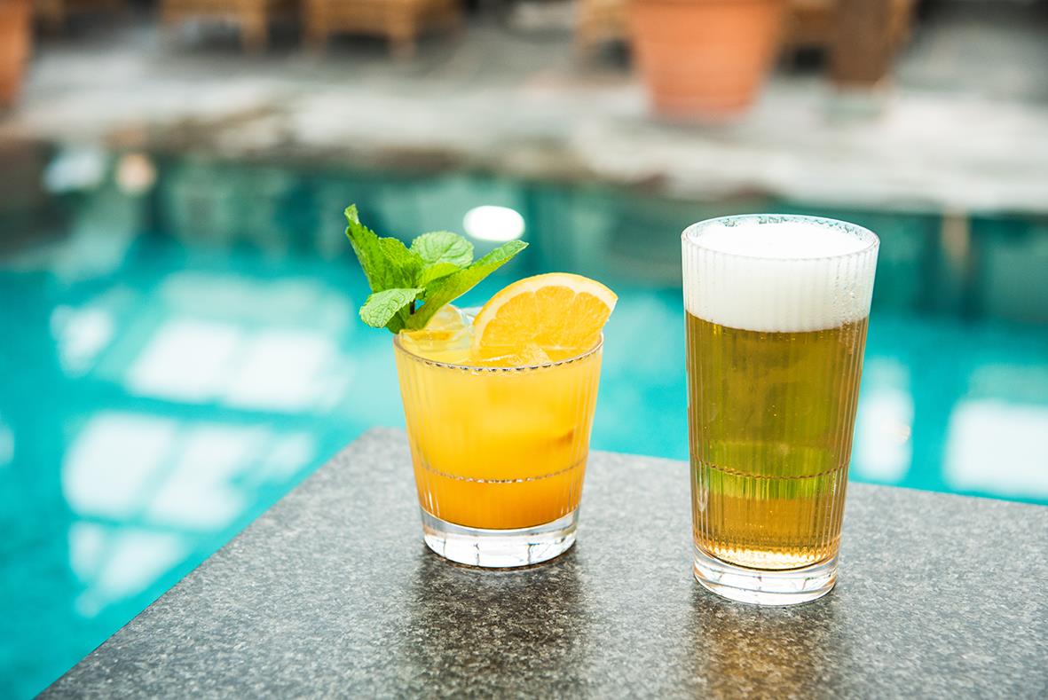 Två drinkglas, ett med orange dryck och mynta, ett med öl, på en poolkant