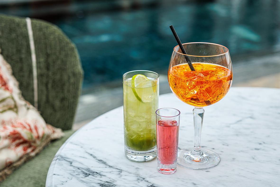 Tre drinkglas på ett bord, ett stort gin & tonicglas och två mindre glas med olika drycker, utomhus vid pool.
