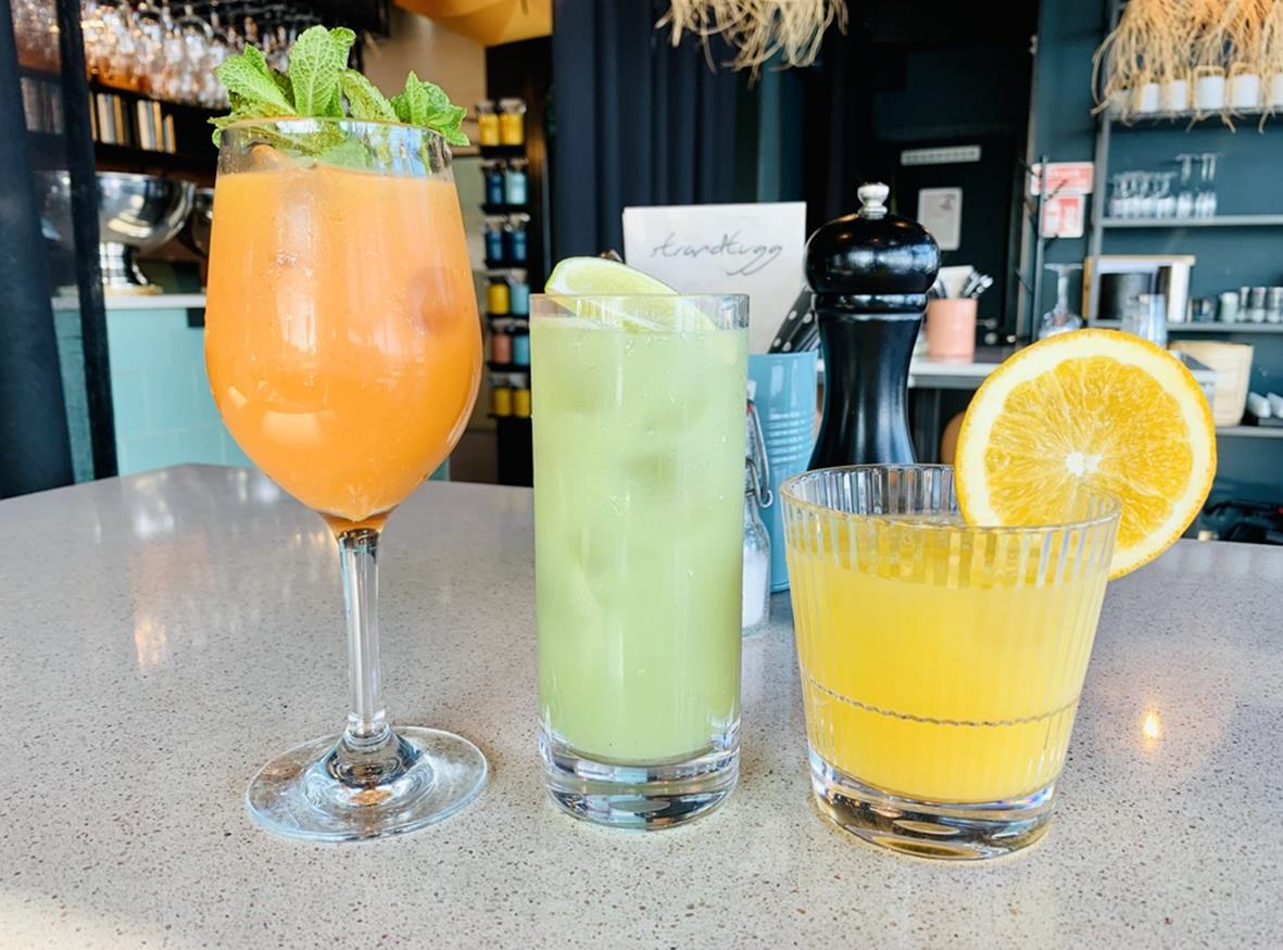 Tre drinkglas med olika drycker på bar eller café, ett med orange drink, ett med grön drink och ett med gul drink och citron.