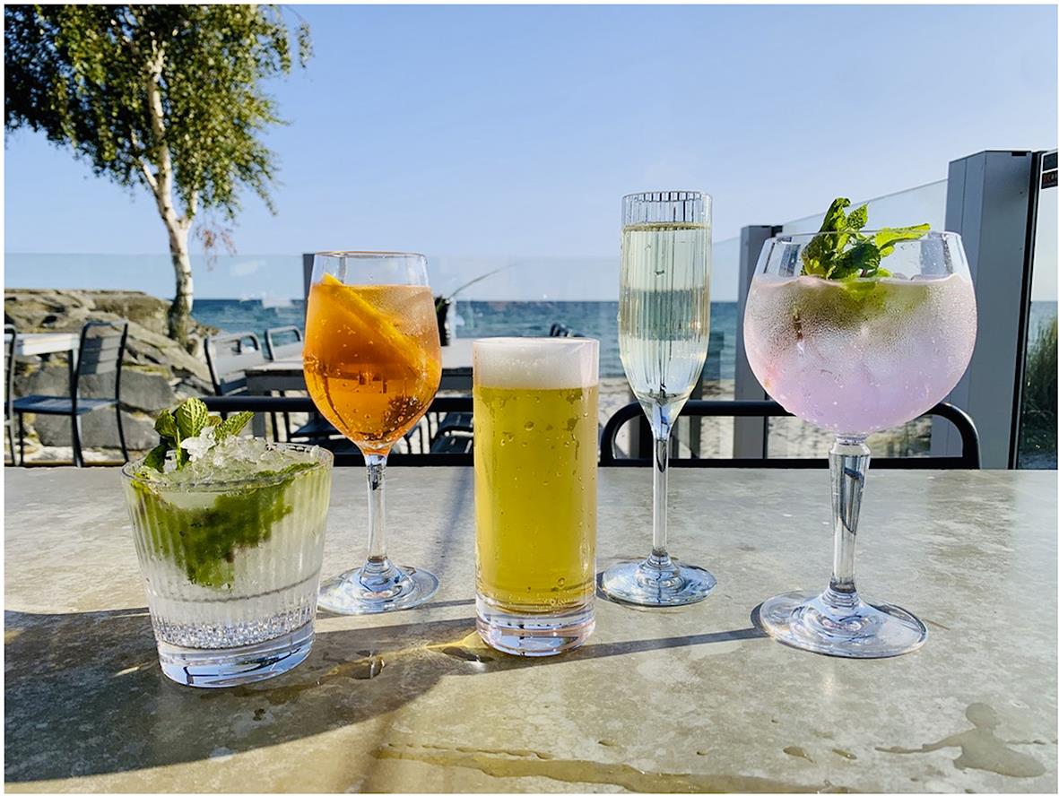 Fyra drinkglas med olika drycker på bord vid havet, bakgrund av träd och klarblå himmel