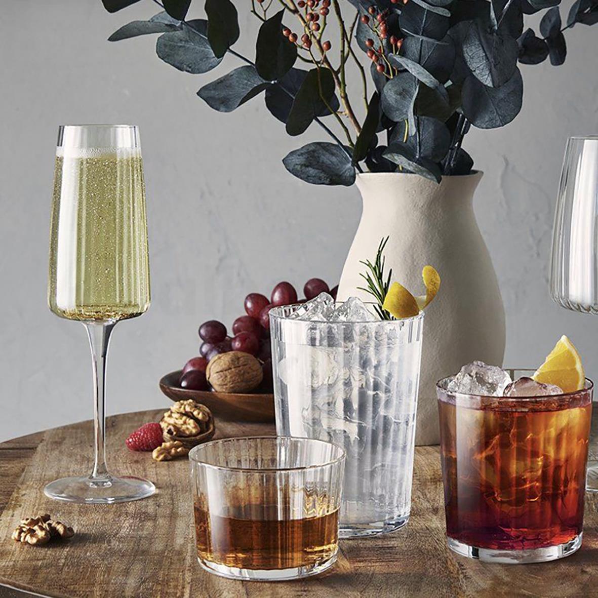 Drinkglas Bormioli Rocco Exclusiva Rum med citrusskivor och is, på träbord med vin och frukt i bakgrunden