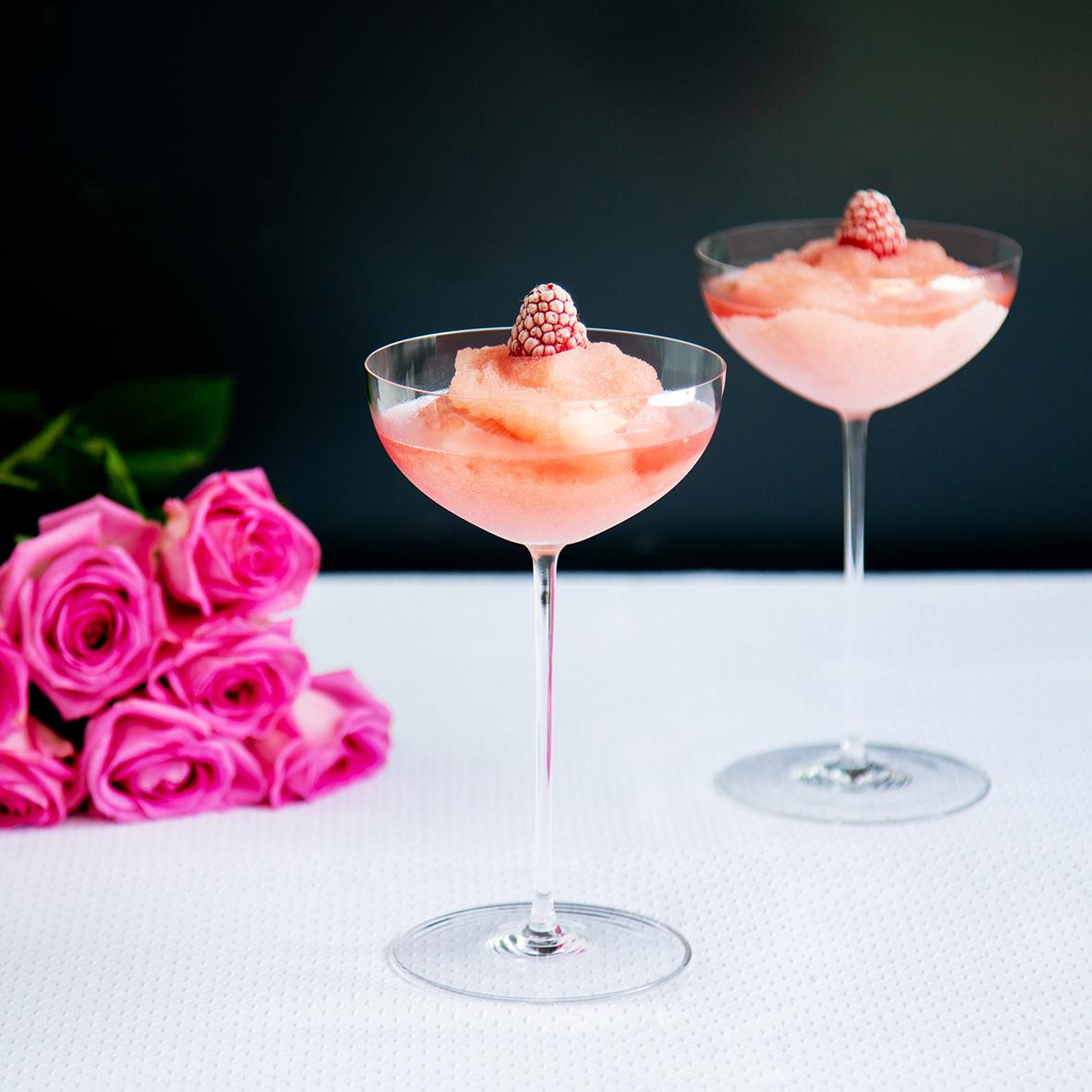 Två cocktailglas med rosa drink och hallon, rosa rosor bredvid, på vit duk