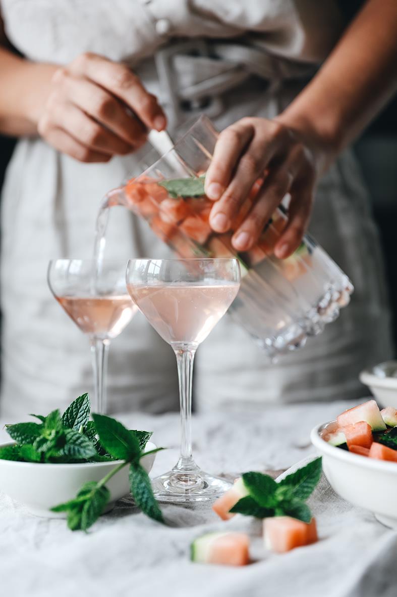 Person häller drink i Riedel cocktailglas med is och mynta, omgivna av frukt och mynta
