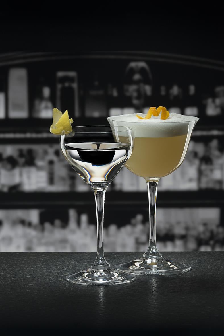 Två cocktailglas, ett med svart drink och ett med vit mousse, på barhylla med suddig bakgrund