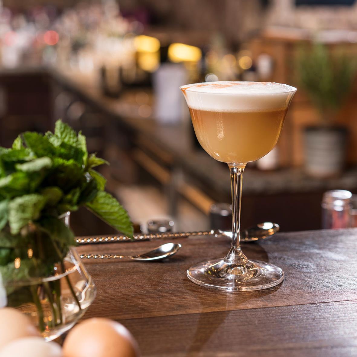 Cocktailglas Riedel Drink Specific Sour med frostad yta på en träbord, bakgrund av barmiljö