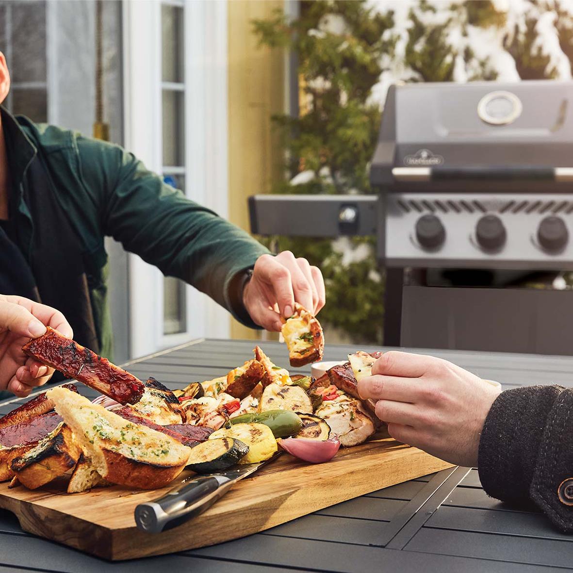 Person som äter grillad mat från en träskärbräda framför en gasolgrill Napoleon Freestyle 365