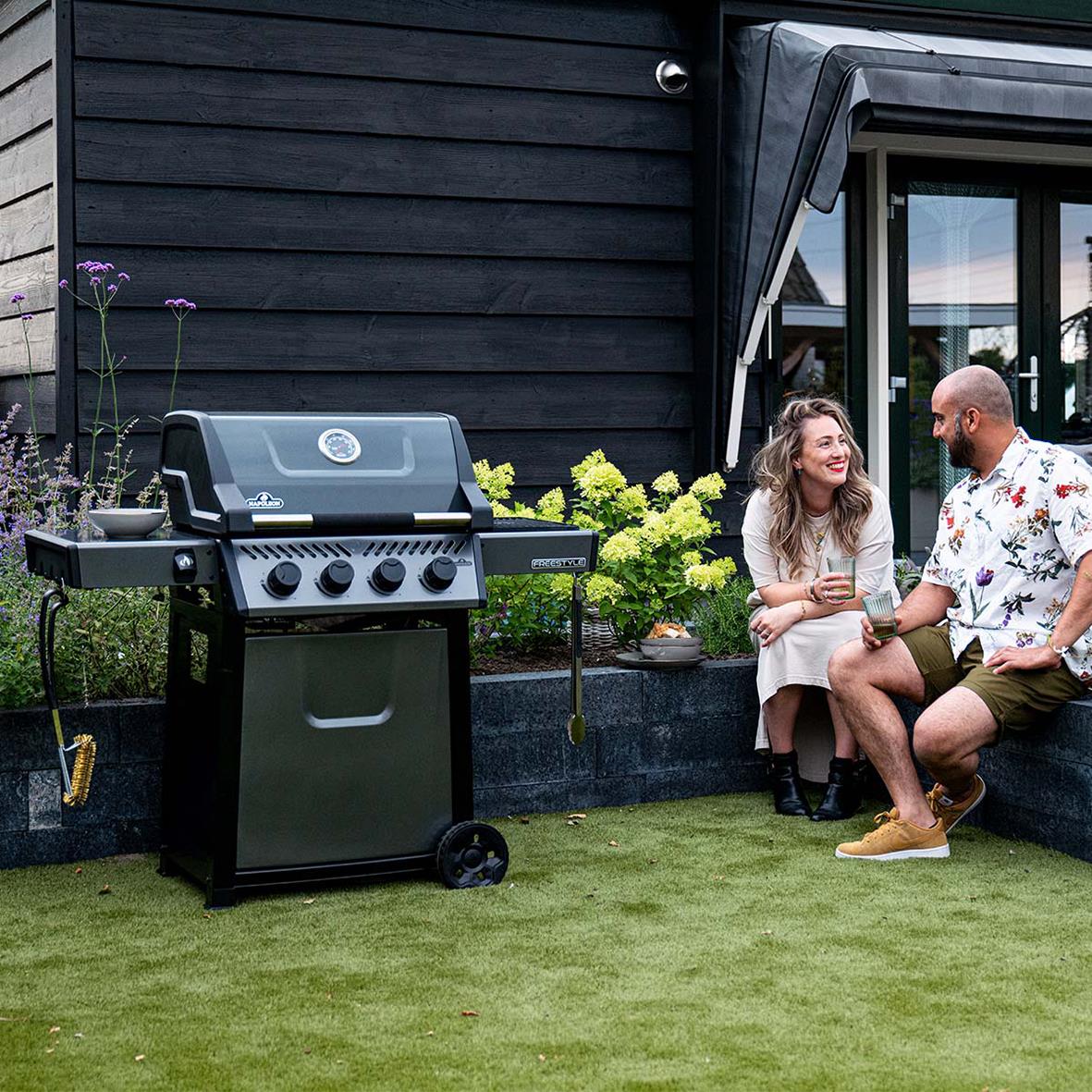 Två personer sitter och pratar bredvid en svart gasolgrill på en gräsmatta utomhus