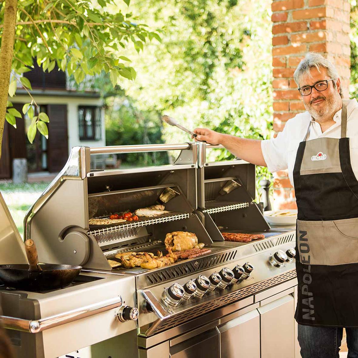 Man grillar på en Napoleon Prestige PRO 825-3 gasolgrill utomhus med grönt träd och hus i bakgrunden