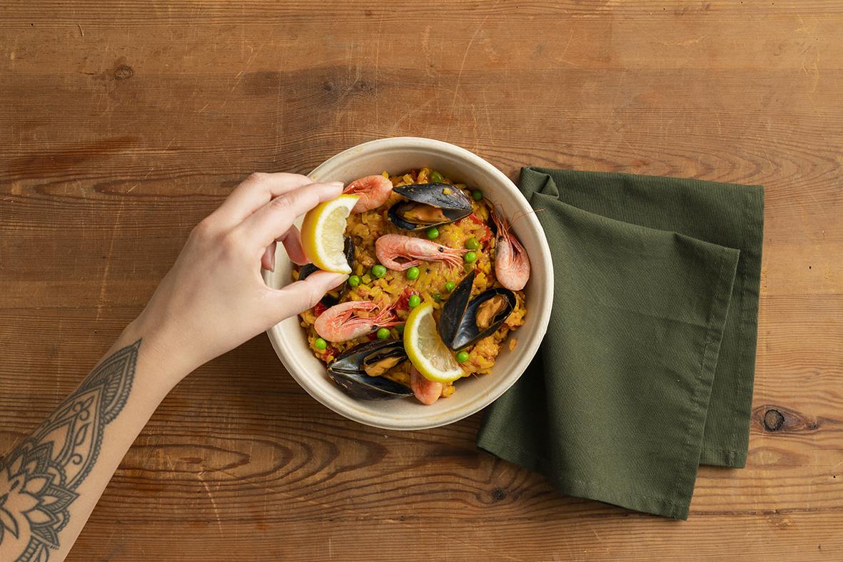 En hand håller en salladsskål med skaldjur och citron, bredvid en mörkgrön servett på ett träbord