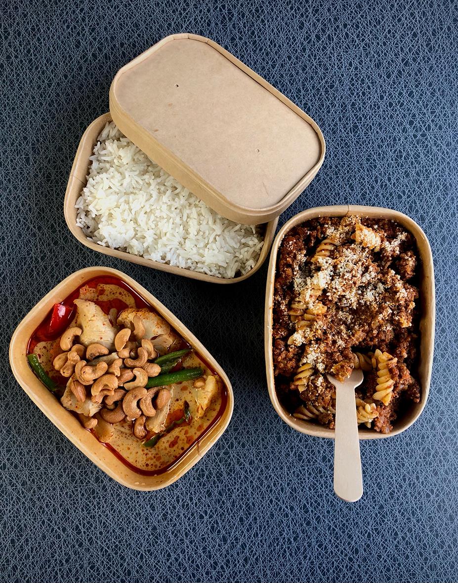 Tre pappersformar i kartong, en med ris, en med köttfärssås och pasta, en med kyckling och grönsaker, på blå bakgrund