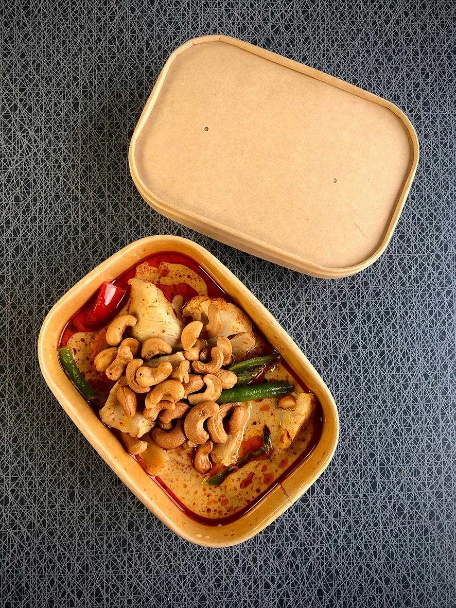 Brun pappersform med kyckling- och cashewnötter i en sås, öppnad lock bredvid på mörk bakgrund