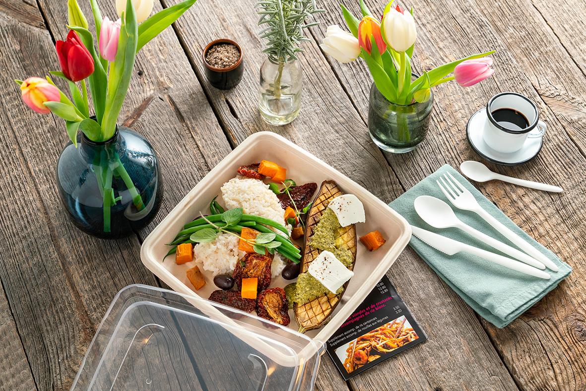 En matlåda med mat, omgiven av tulpaner i vaser, bestick och en kopp kaffe på ett träbord