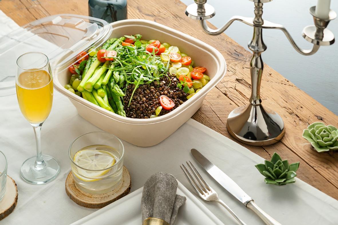 Stor sallad i en beige bagasse-kopp på ett bord med glas, bestick och ljusstake