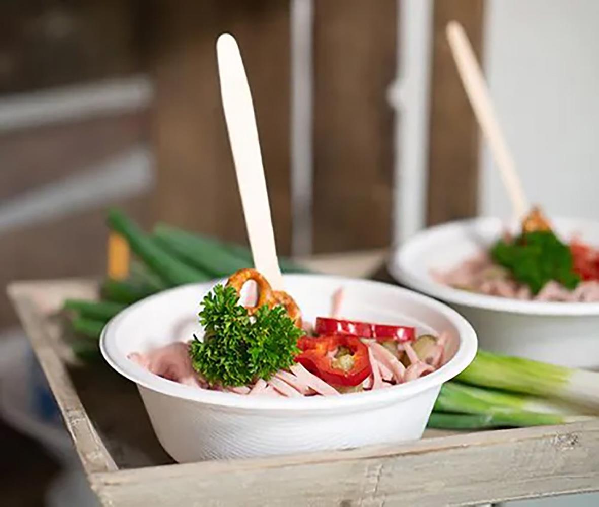 Vit rund skål av bagasse med sallad, grönsaker och en sked, på träbricka