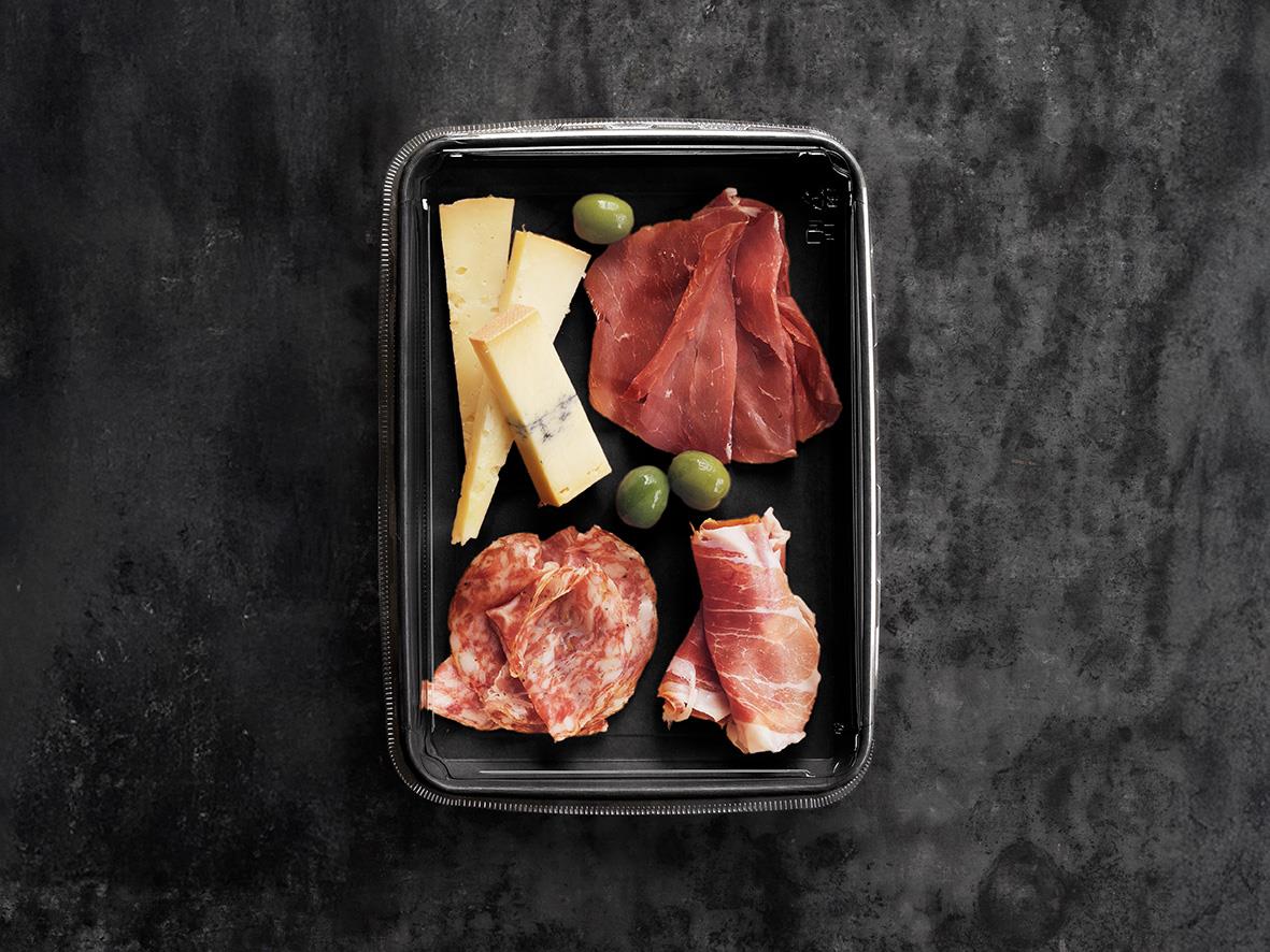 Svart pappersform med ost, skinka, salami och gröna oliver på mörk bakgrund