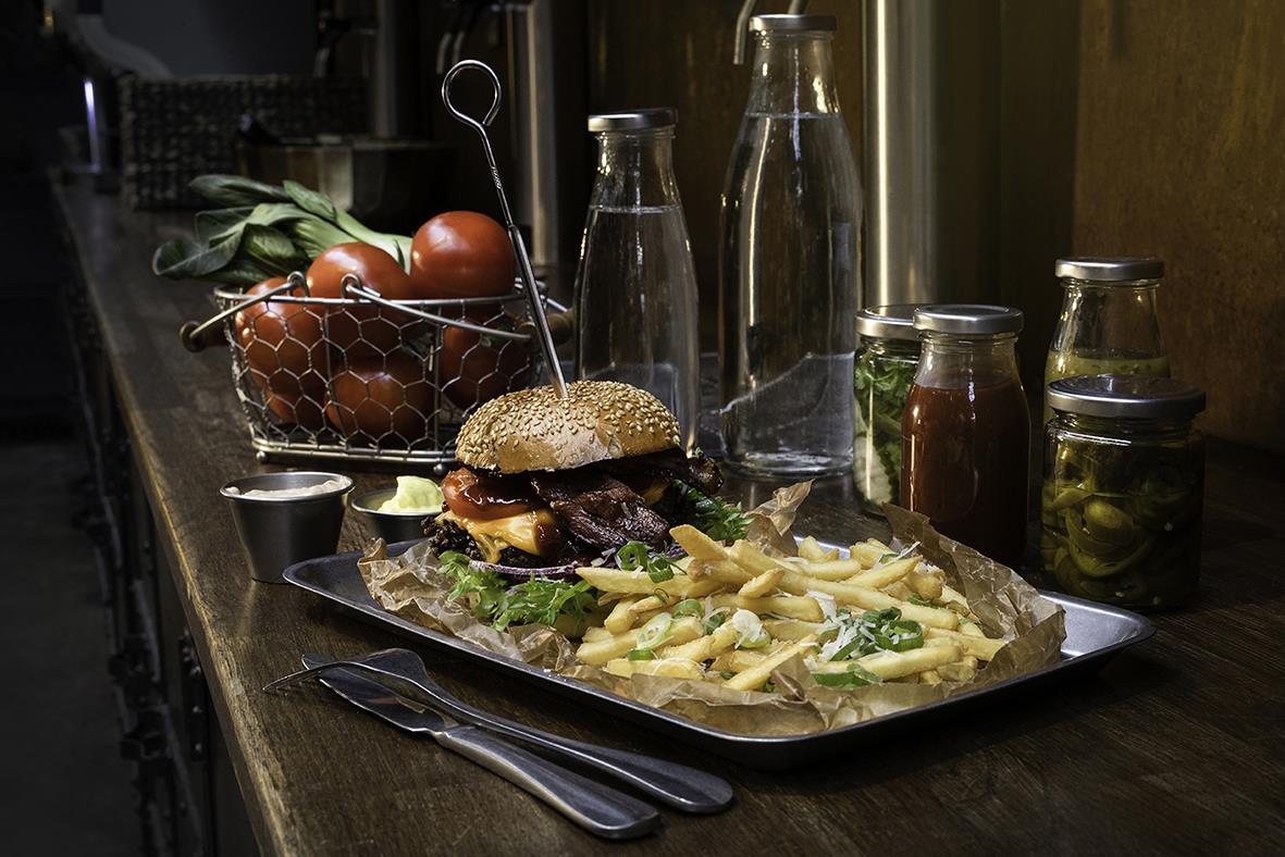 Serveringsbricka med hamburgare, pommes frites, grönsaker, flaskor och burkar på ett träbord