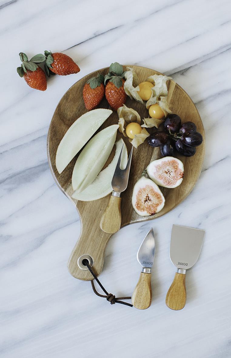 Ostbricka med jordgubbar, melon, physalis och druvor på rund träbricka med ostknivar på marmorbord