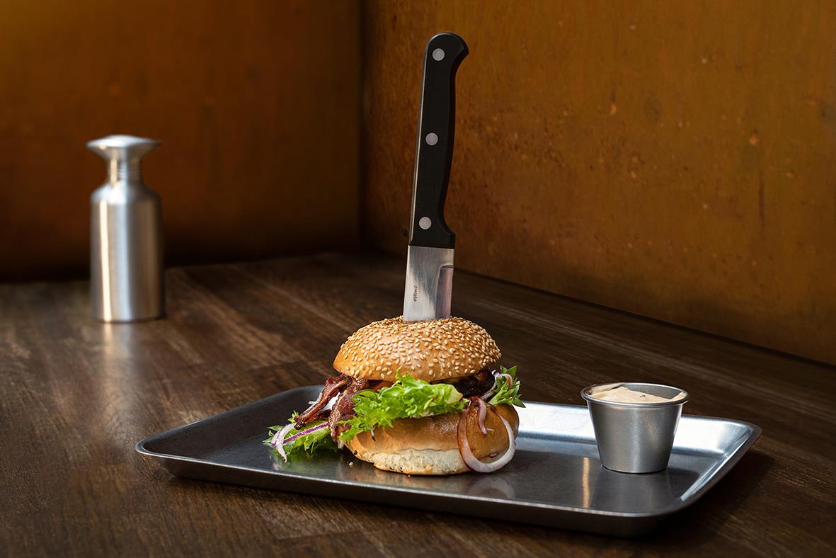 Dressingkopp i rostfritt stål på en bricka med en kniv, en hamburgare och en liten kanna