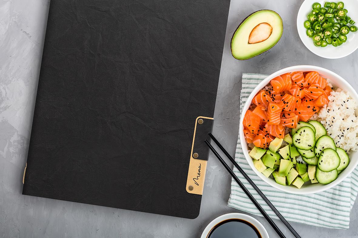 Svart menypärm i A4 storlek med guldklämma, omgiven av avokado, sallad och sushi