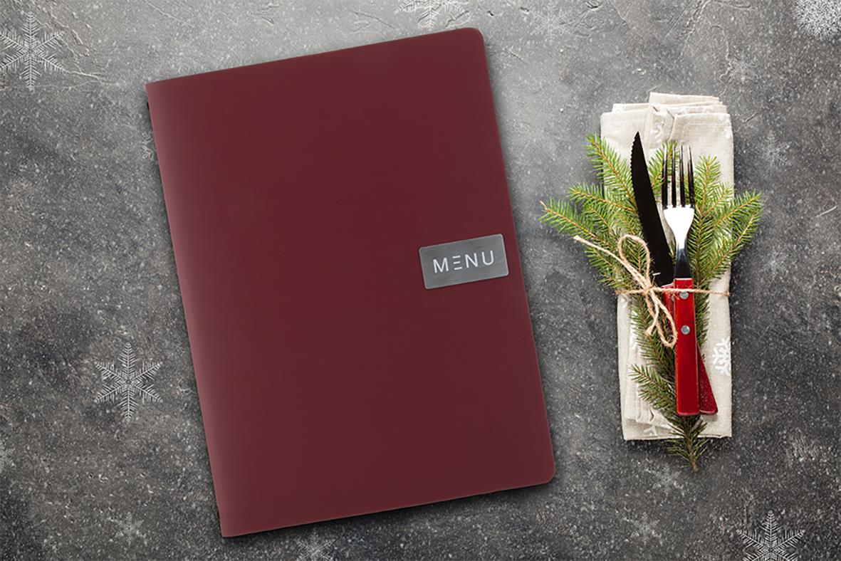 Röd menypärm i A4 storlek med etikett Meny på framsidan, placerad på en mörk yta bredvid en servett med kniv, gaffel och grön kvist