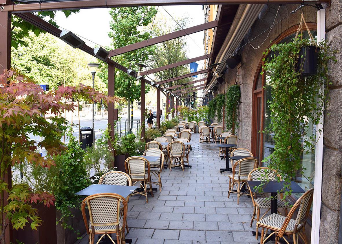 " med bord och stolar längs en gata med växter och träd, under ett tak"