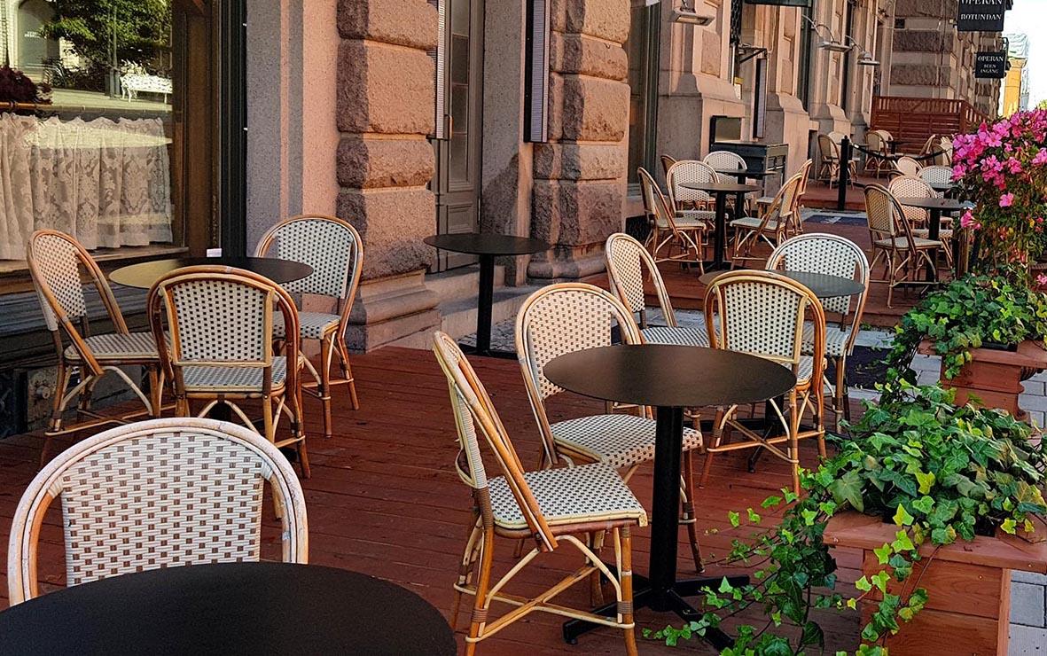 Utomhusbord med svart bordskiva och vita stolar med rottingdetaljer på uteservering