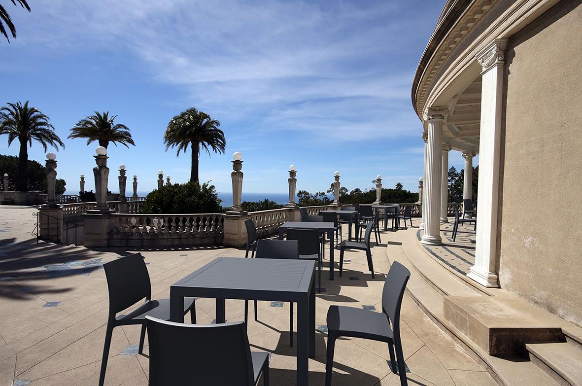 Uteservering med svarta cafébord och stolar på en terrass med utsikt över havet och palmer