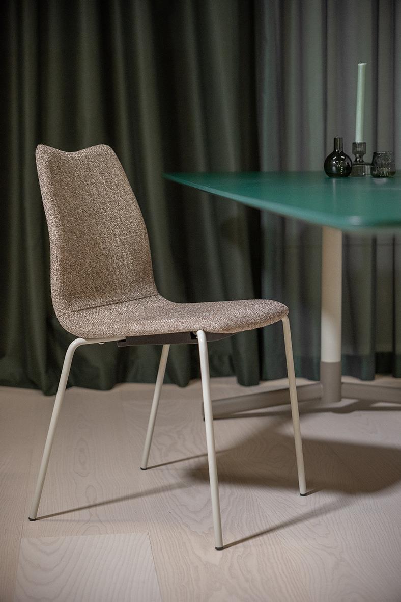 En modern stol med grått tygsäte och vita metallben framför ett grönt bord med dekorationer och mörk gardin i bakgrunden