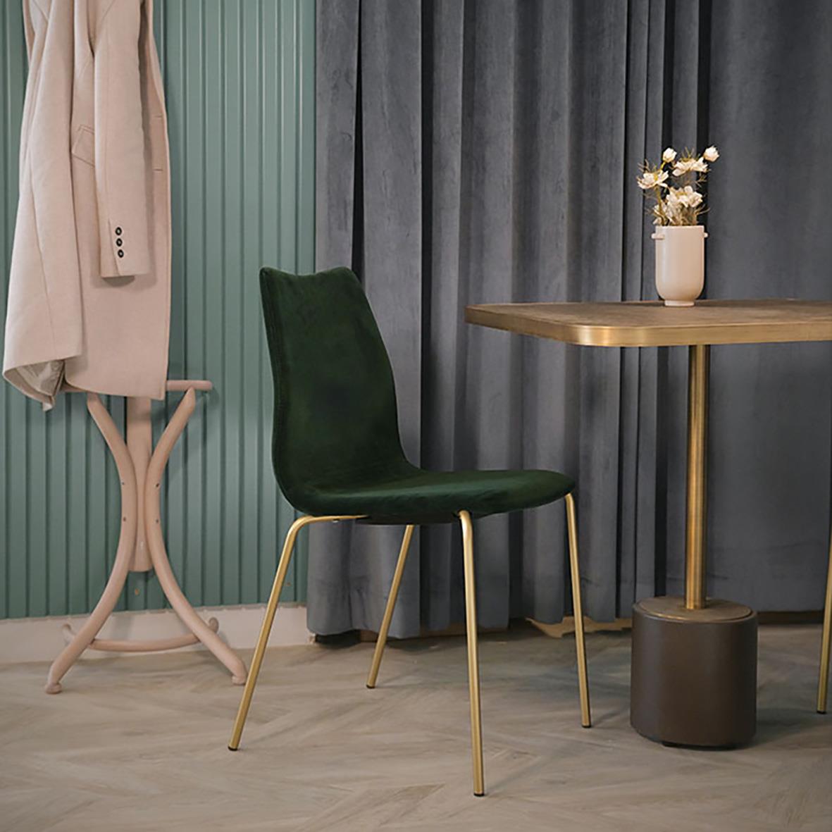 Mörkgrön stol vid ett runt bord med blomsterarrangemang, bakgrund av gröna och mörkgråa gardiner.