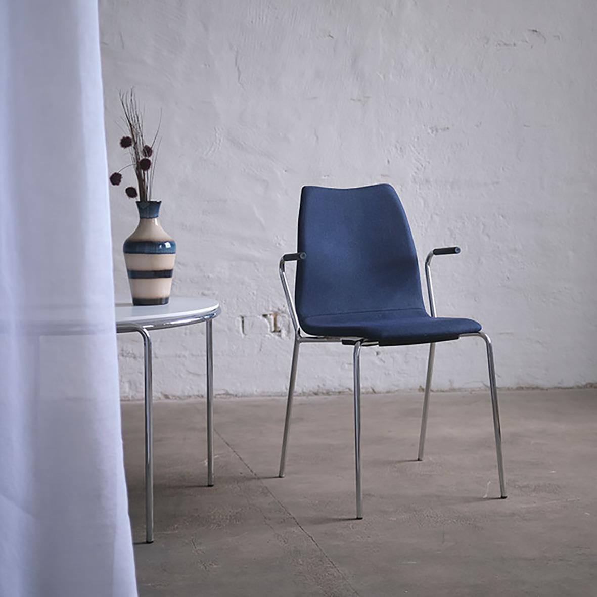 En blå stol bredvid ett runt bord med en vas och blommor mot en vit vägg