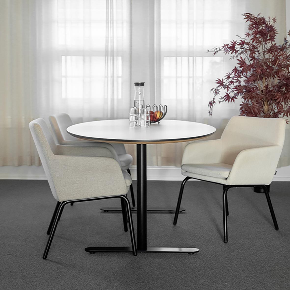 Runt bord med fyra beige stolar och en vit armstol, bakom fönster med vita gardiner och en växt