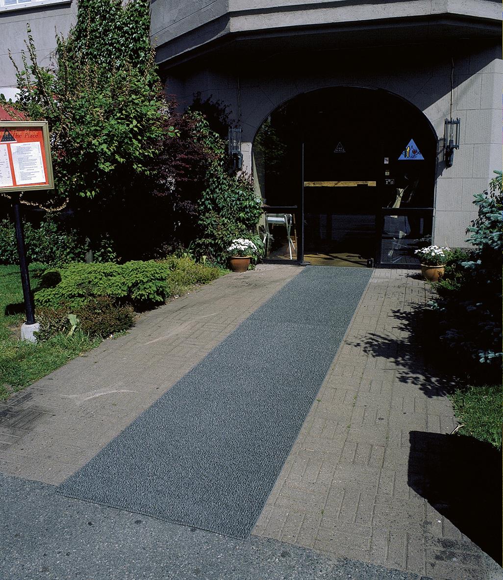 Skrapmatta Astro Turf 55x90cm framför en ingång under en båge, omgiven av gröna buskar och träd