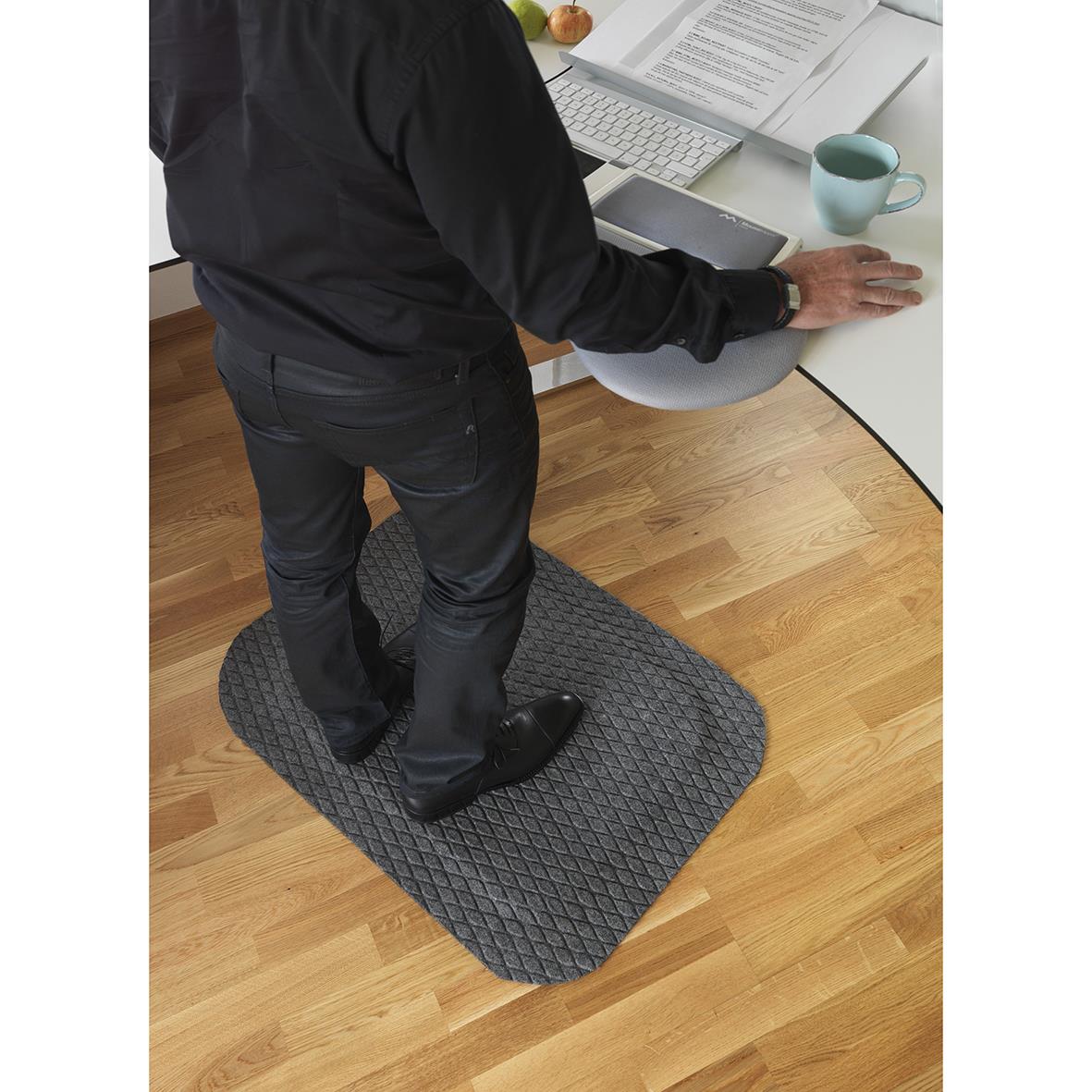 Person står på en arbetsplatsmatta framför ett skrivbord med dokument, dator och kopp på.