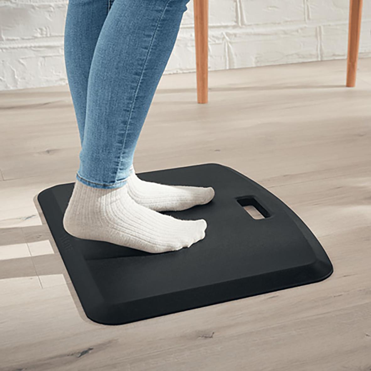 Person står på en mörkgrå ergonomisk ståmatta Leitz Ergo Cosy i ett hem, med vita sockor och jeans, på ett trägolv.