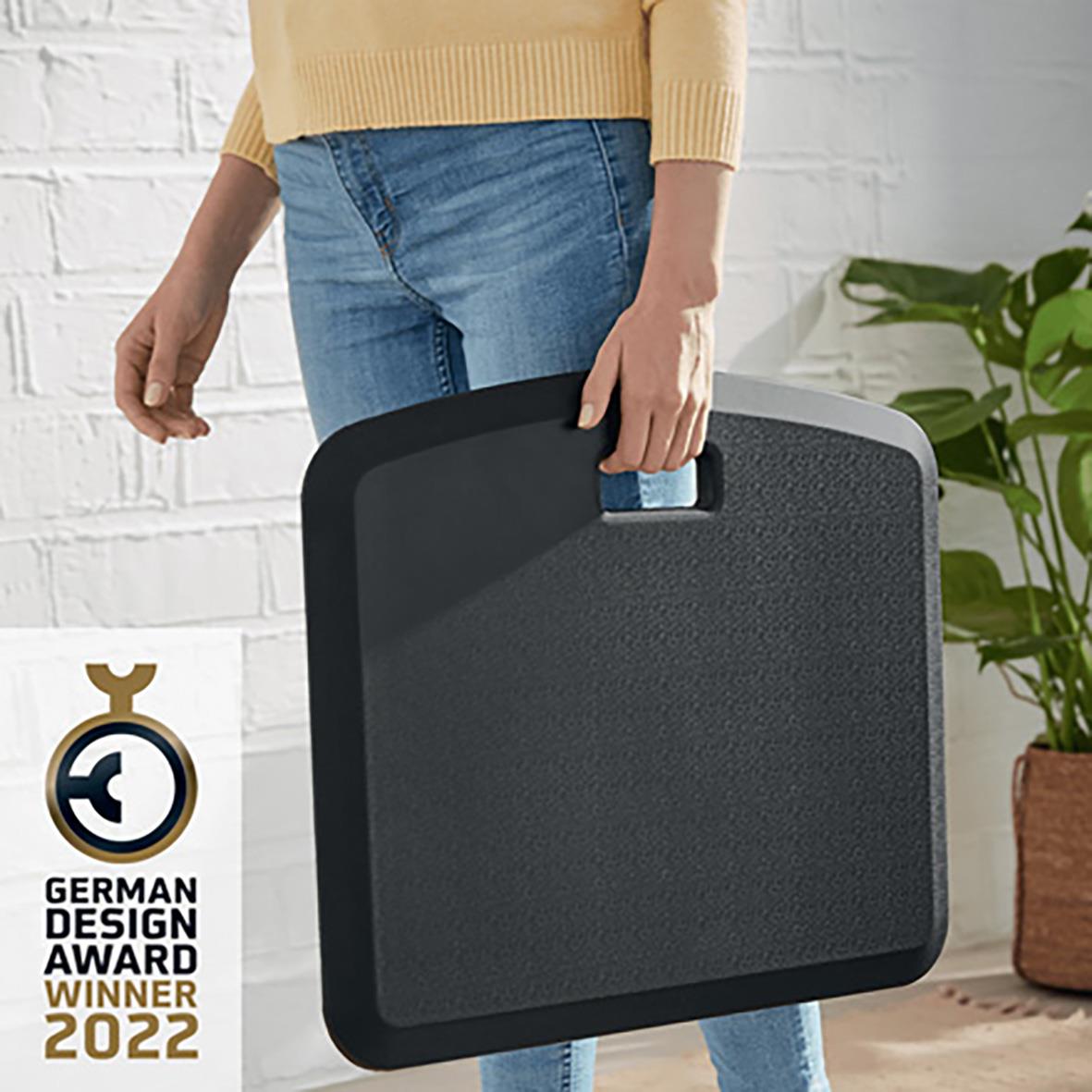 Person håller en mörkgrå ergonomisk ståmatta Leitz Ergo Cosy, designad för komfort, i ett inomhusmiljö med växt i bakgrunden
