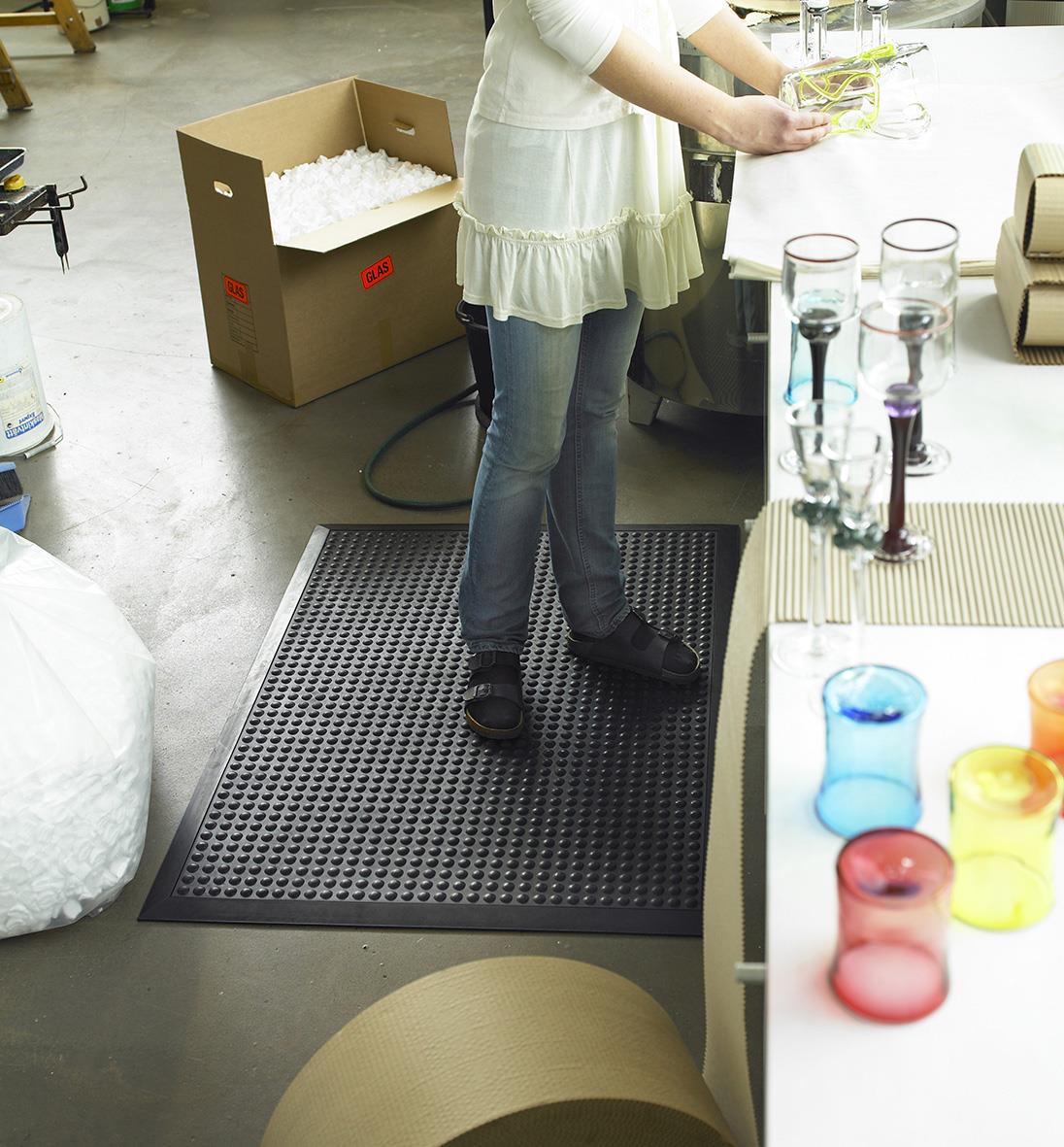 Person står på en svart arbetsplatsmatta i ett kontorsmiljö med glas och andra föremål runt omkring