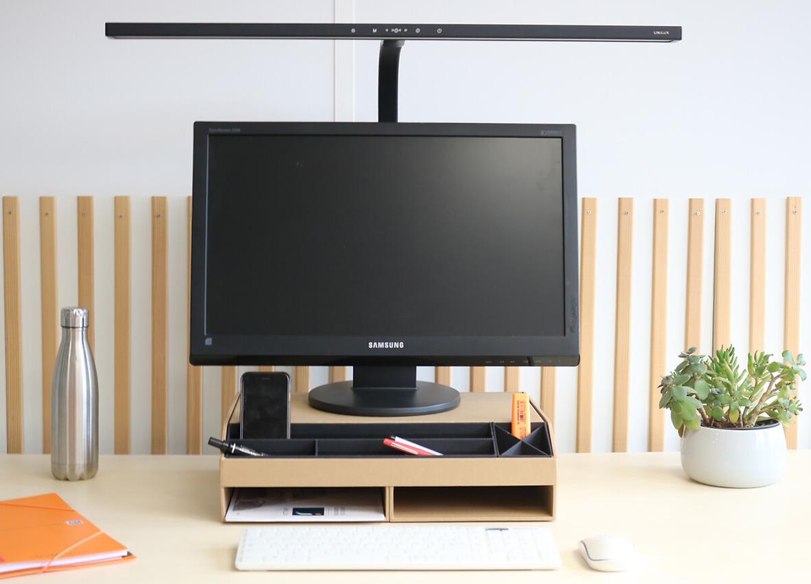 Skrivbord med svart datorskärm, Unilux Strata Lumino lampa, vätskedunk, växt och skrivmaterial
