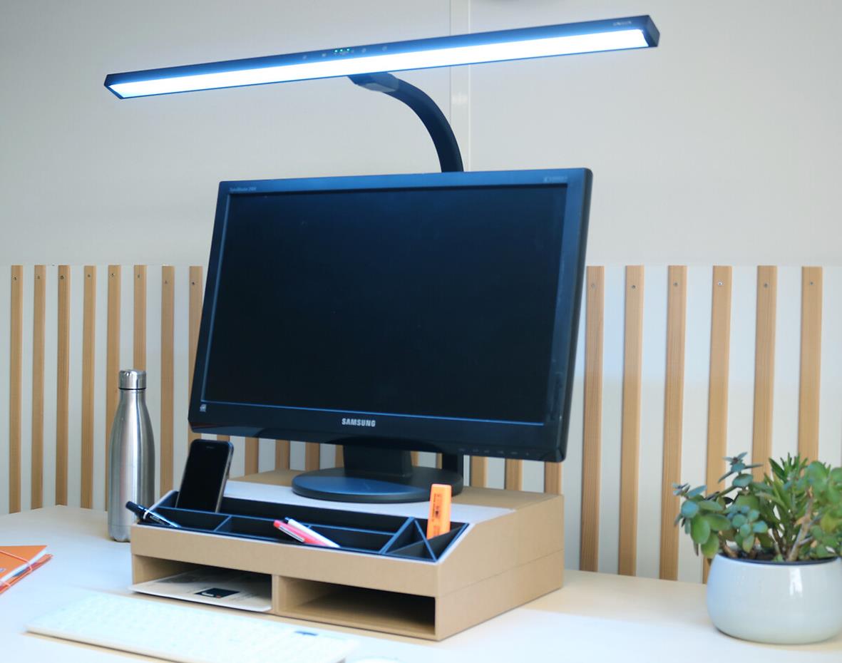 Skrivbordslampa Unilux Strata Lumino svart ovanför dator på skrivbord med växt och vattenflaska