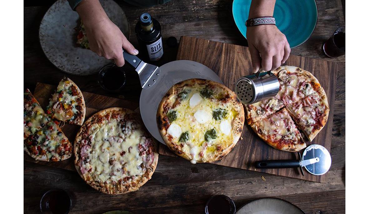 Pizza server, skärare och kryddströare på bord med pizzor och händer som skär och serverar