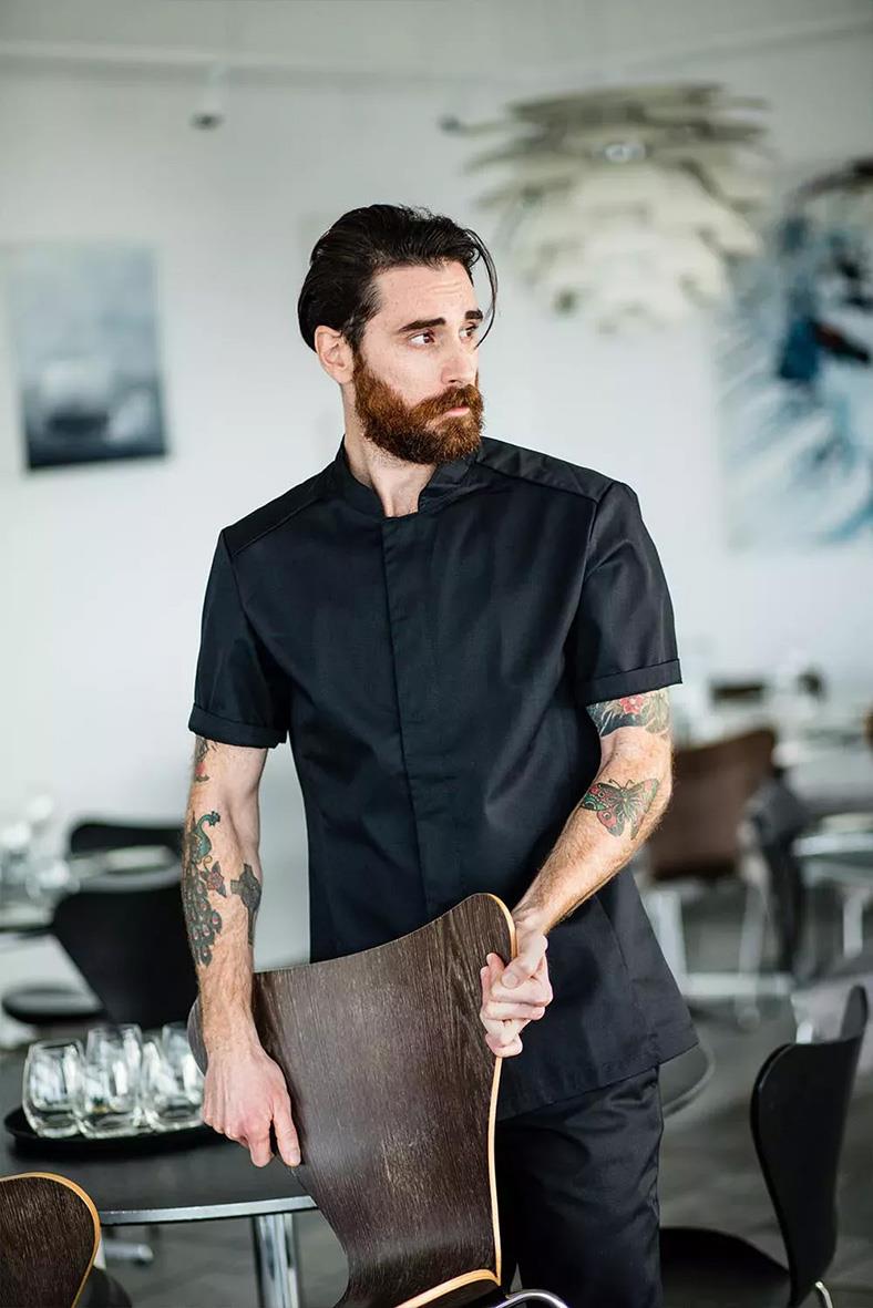 Man i svart kockskjorta i ett modernt restaurangkök, håller en stol, med tatueringar på armarna