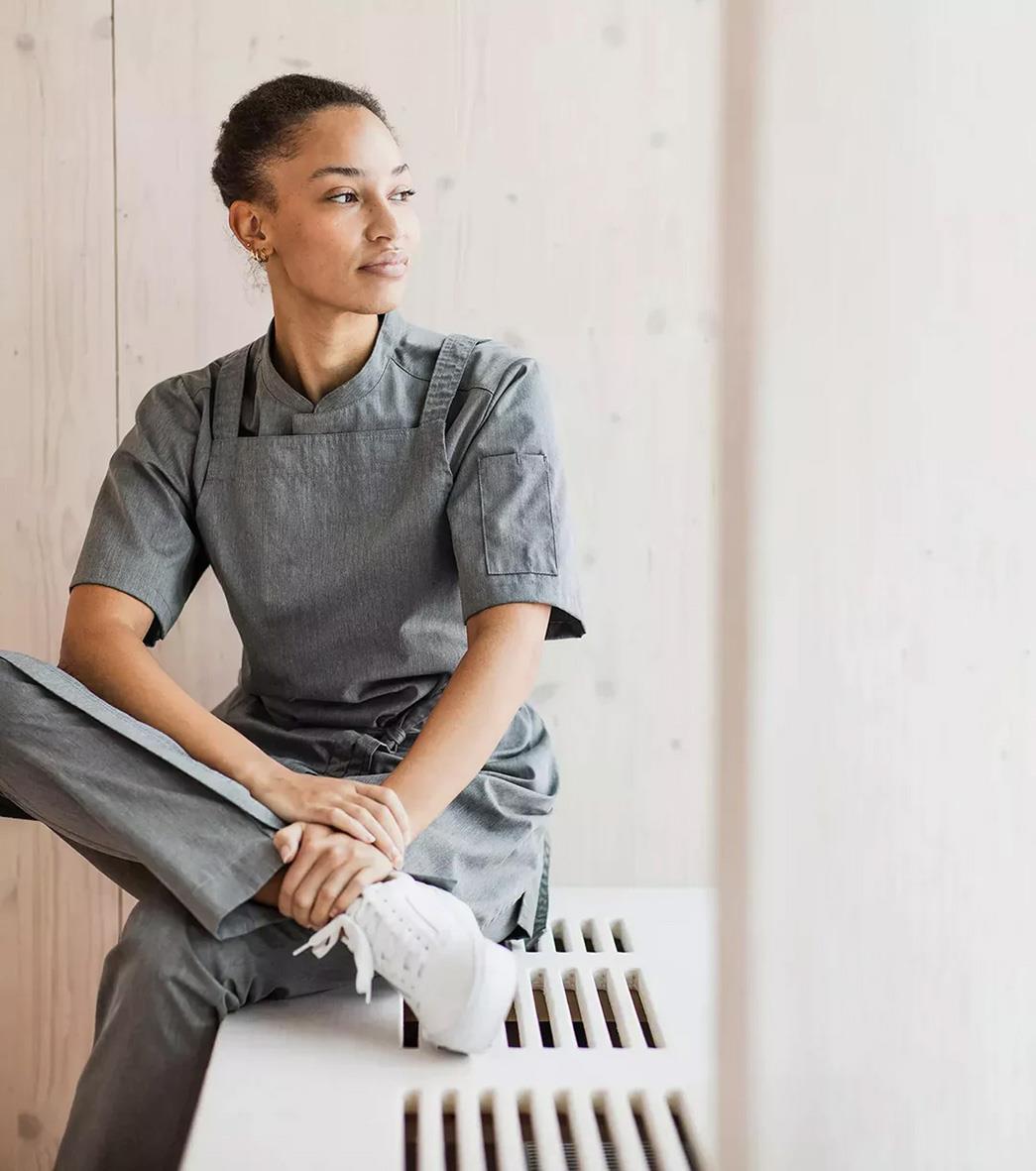 Ung kvinna i grå melange kockskjorta och byxor, sittande på en bänk, ser åt sidan