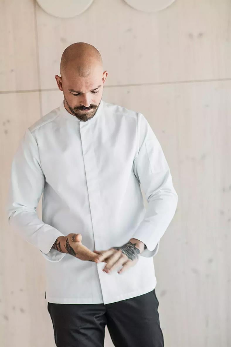 Man i vit kockskjorta med svart byxa, står mot ljus vägg